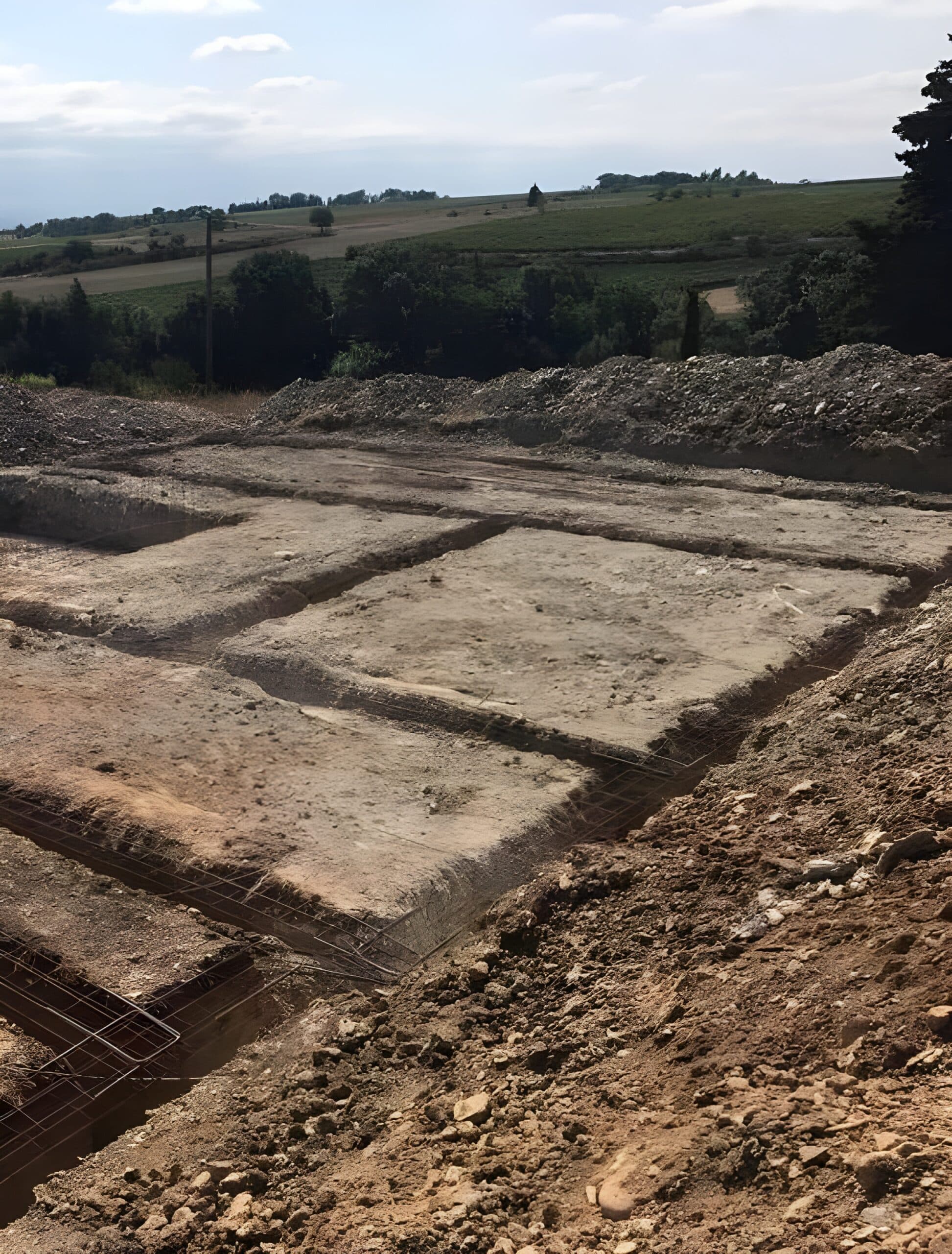 Tranchées de fondation préparées avec ferraillage sur un terrain en pente. Vue sur les tas de terre excavée et le paysage verdoyant.